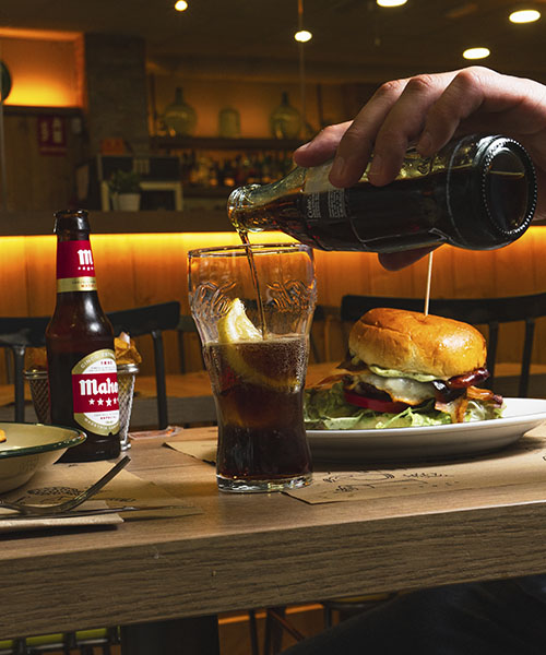 Fotografía de mesa con hamburguesa, cerveza Mahou y refresco servido, realizada por La Pecera para campañas visuales en entornos de restauración.
